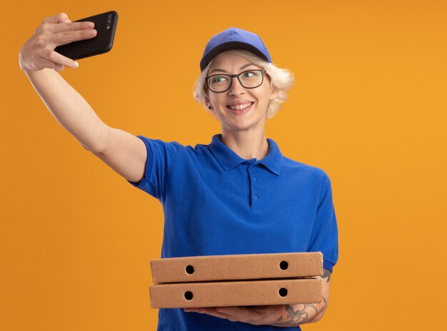 Młoda Kobieta Dostawy W Niebieskim Mundurze I Czapce W Okularach, Trzymając Pudełka Po Pizzy Za Pomocą Smartfona, Robi Selfie, Uśmiechając Się Nad Pomarańczową ścianą