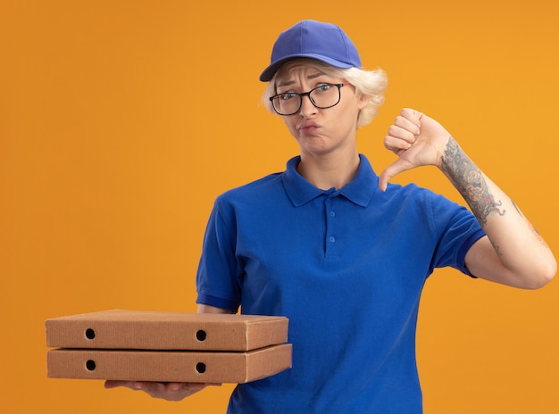 Młoda kobieta dostawy w niebieskim mundurze i czapce w okularach trzymająca pudełka po pizzy niezadowolona, pokazując kciuki w dół na pomarańczowej ścianie