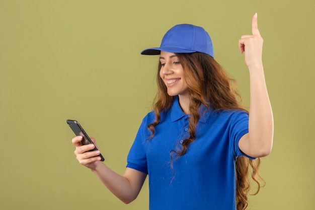 Młoda Kobieta Dostawy Z Kręconymi Włosami Na Sobie Niebieską Koszulkę Polo I Czapkę Patrząc Na Smartfona Uśmiechnięty Palec Wskazujący W Górę Na Odosobnionym Zielonym Tle