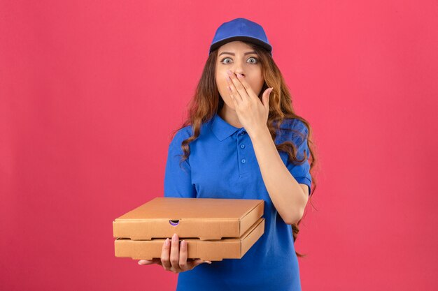 Młoda kobieta dostawy z kręconymi włosami na sobie niebieską koszulkę polo i czapkę stojącą z pudełkami po pizzy zaskoczona zakrywając usta ręką na białym tle różowym