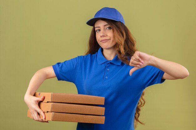 Młoda kobieta dostawy z kręconymi włosami na sobie niebieską koszulkę polo i czapkę stojącą ze stosem pudełek po pizzy pokazującą kciuk w dół pokazujący niechęć na odosobnionym zielonym tle