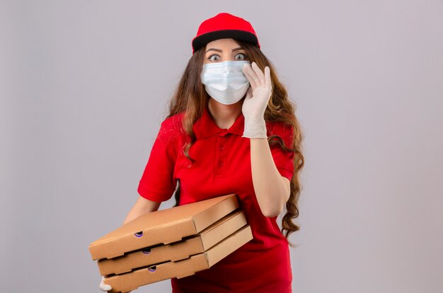 Młoda kobieta dostawy z kręconymi włosami, ubrana w czerwoną koszulkę polo i czapkę w medycznej masce ochronnej i rękawiczkach, stojąca z pudełkami po pizzy, patrząc zaskoczona ręką w pobliżu ust, mówiącą o tajemnicy
