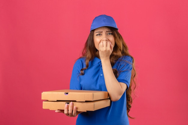 Młoda kobieta dostawy z kręconymi włosami, ubrana w niebieską koszulkę polo i czapkę stojącą z pudełkami po pizzy, wyglądająca na zestresowaną i zdenerwowaną, z rękami na ustach gryzącymi paznokcie na izolowanym różowym tle