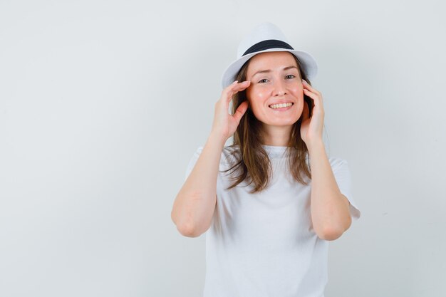 Młoda kobieta dotyka skóry twarzy na policzkach w białej koszulce, kapeluszu i ładnie wygląda.