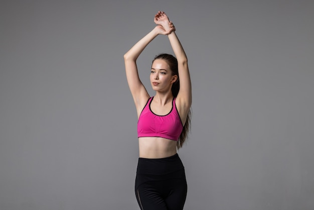 Młoda kobieta fitness lekkoatletycznego w odzieży sportowej, rozciągając ramiona i patrząc na kamery, stojąc na szarym tle. Koncepcja zdrowego stylu życia i sportu
