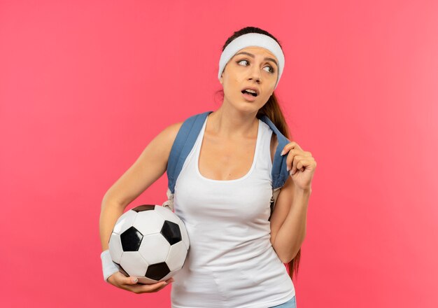 Młoda kobieta fitness w odzieży sportowej z opaską i złotym medalem na szyi z plecakiem trzymającym piłkę nożną patrząc na bok zdziwiony stojąc nad różową ścianą