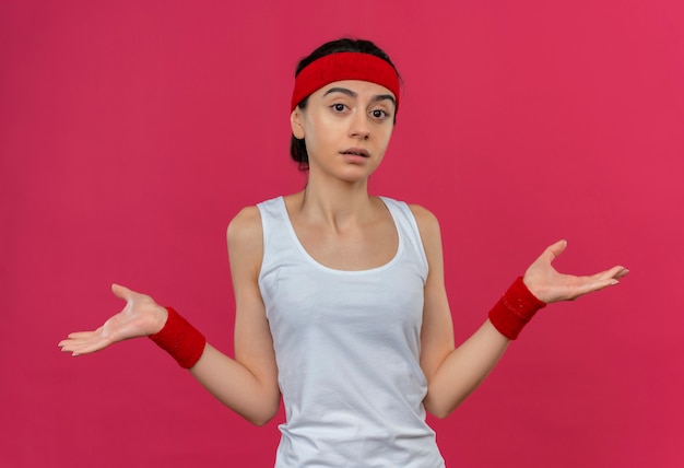 Młoda kobieta fitness w odzieży sportowej z opaską na głowę wyglądająca na zdezorientowaną i niepewną, rozkładającą ręce na boki, nie mając odpowiedzi, stojącą nad różową ścianą