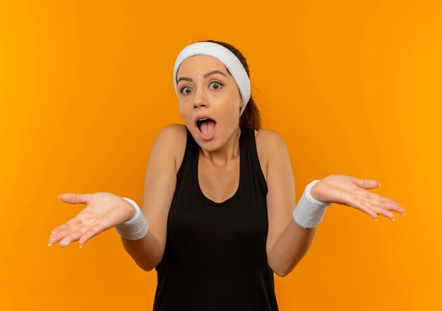 Młoda kobieta fitness w odzieży sportowej z pałąkiem na głowę, patrząc zaskoczony i zmieszany, wzruszając ramionami, stojąc na pomarańczowej ścianie