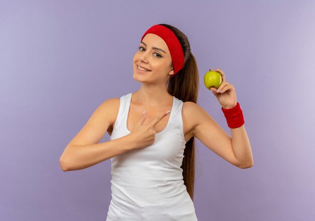 Młoda kobieta fitness w odzieży sportowej z pałąkiem na głowę trzyma zielone jabłko, wskazując palcem na to, uśmiechając się pewnie stojąc na szarej ścianie