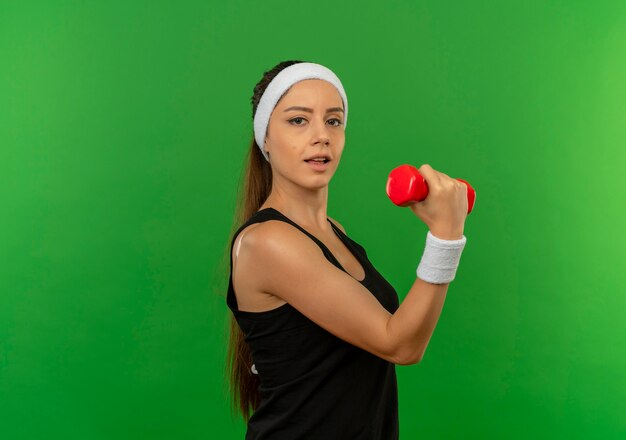 Młoda kobieta fitness w odzieży sportowej z pałąkiem na głowę, trzymając hantle, robi ćwiczenia patrząc pewnie stojąc nad zieloną ścianą