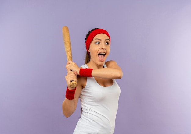 Młoda kobieta fitness w odzieży sportowej z pałąkiem na głowę, trzymając kij bejsbolowy grając szczęśliwy i podekscytowany stojąc na szarej ścianie