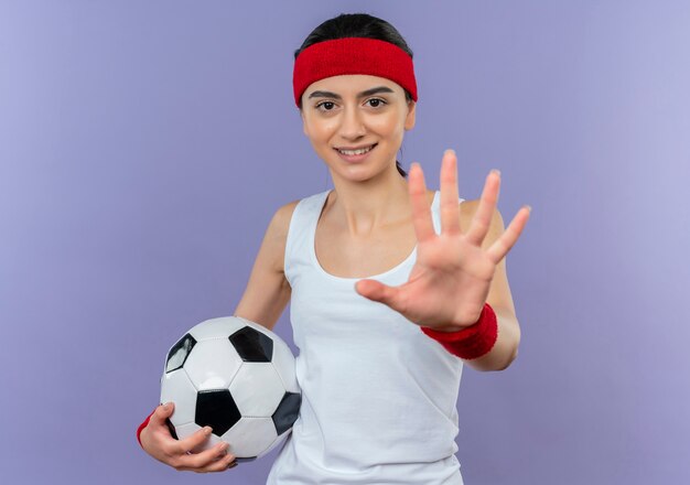 Młoda kobieta fitness w odzieży sportowej z pałąkiem na głowę, trzymając piłkę nożną co znak stopu z otwartą dłonią stojącą nad fioletową ścianą