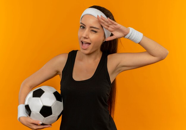 Młoda kobieta fitness w odzieży sportowej z pałąkiem na głowę, trzymając piłkę nożną mrugając i uśmiechając się, salutując stojąc nad pomarańczową ścianą