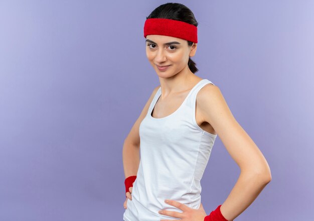 Młoda kobieta fitness w odzieży sportowej z pałąkiem na głowę z pewnym uśmiechem stojącym nad fioletową ścianą