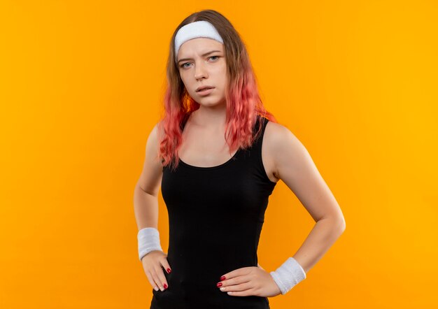 Młoda kobieta fitness w odzieży sportowej z pewną poważną miną stojącą nad pomarańczową ścianą