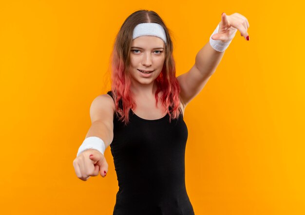 Bezpłatne zdjęcie młoda kobieta fitness w odzieży sportowej z pewnym siebie uśmiechem wskazując palcami na aparat stojący nad pomarańczową ścianą
