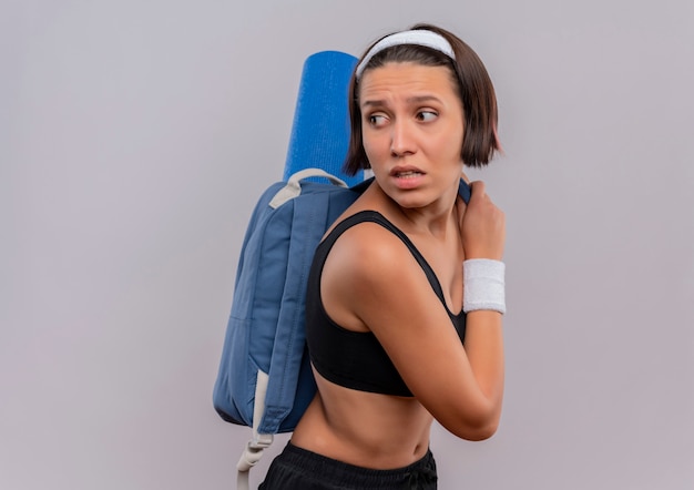 Bezpłatne zdjęcie młoda kobieta fitness w odzieży sportowej z plecakiem i matą do jogi, patrząc wstecz z wyrazem strachu stojąc na białej ścianie