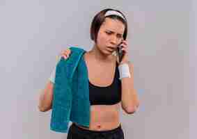 Bezpłatne zdjęcie młoda kobieta fitness w odzieży sportowej z ręcznikiem na ramieniu, patrząc zdezorientowany i niespokojny podczas rozmowy na telefon komórkowy stojący nad białą ścianą
