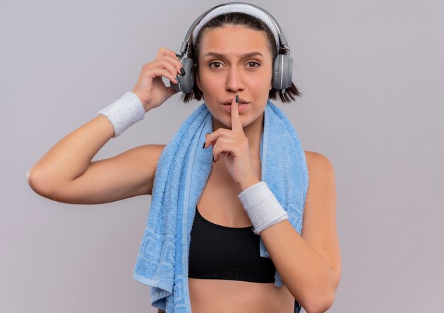 Młoda kobieta fitness w odzieży sportowej ze słuchawkami na głowie i ręcznikiem na szyi, czyniąc gest ciszy z palcem na ustach stojąc na białej ścianie