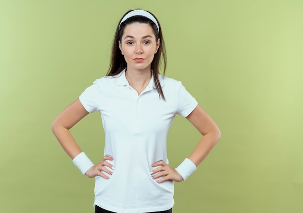 Bezpłatne zdjęcie młoda kobieta fitness w opasce patrząc na kamery z wyrazem pewności z rękami na biodrze, stojąc na jasnym tle