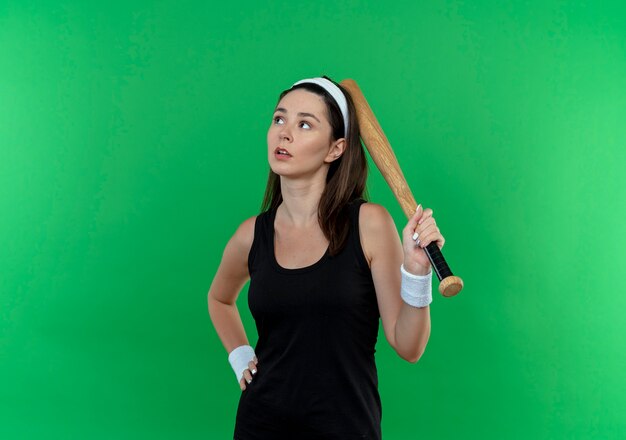 młoda kobieta fitness w opasce trzyma kij baseballowy loking na bok z zamyślonym wyrazem stojącym nad zieloną ścianą