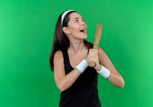 młoda kobieta fitness w opasce trzyma kij bejsbolowy patrząc na bok szczęśliwy i podekscytowany stojąc nad zieloną ścianą