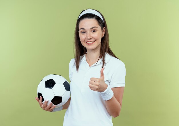 młoda kobieta fitness w opasce trzyma piłkę nożną uśmiechnięty pokazując kciuk do góry stojąc nad ścianą światła