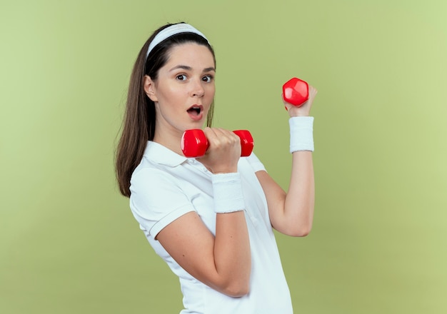 Młoda Kobieta Fitness W Opasce Z Hantlami Patrząc Zaskoczony Stojąc Na Jasnym Tle