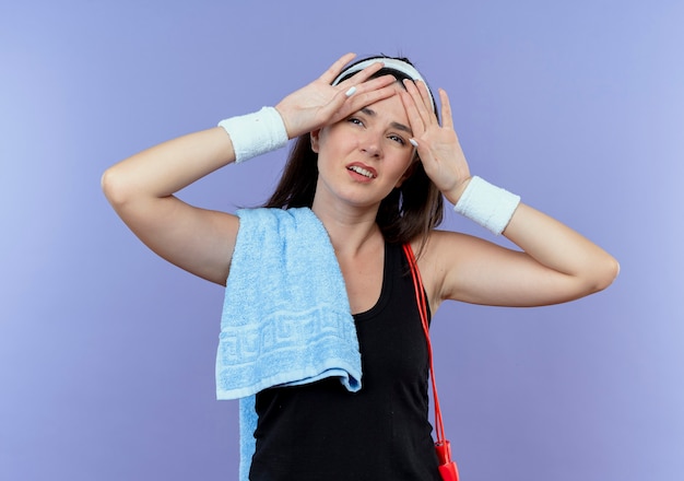 Młoda kobieta fitness w opasce z ręcznikiem na ramieniu, patrząc zmęczony i wyczerpany po treningu stojąc na niebieskim tle