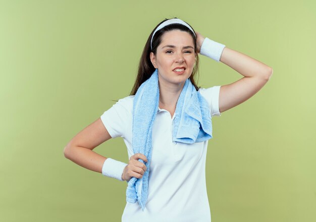 Młoda Kobieta Fitness W Opasce Z Ręcznikiem Na Szyi Patrząc Na Kamery Mylić Z Ręką Na Głowie Za Błąd Stojąc Na Jasnym Tle