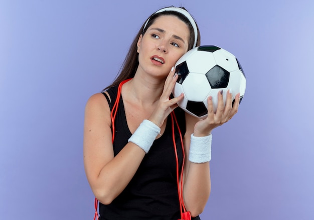 Młoda kobieta fitness w opasce z skakanka wokół szyi trzymając piłkę nożną patrząc na bok opony i znudzony stojący na niebieskim tle