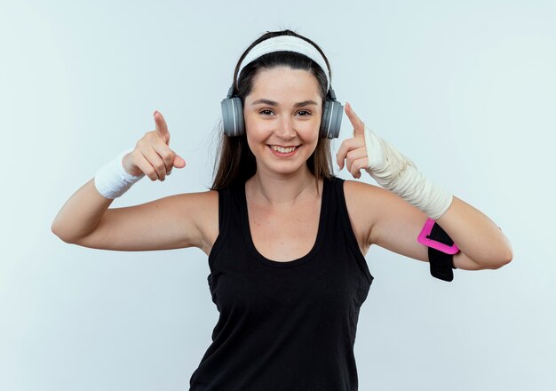 młoda kobieta fitness w opasce ze słuchawkami i opaską na smartfona, uśmiechając się z radosną buźką, wskazując palcami wskazującymi na bok stojący nad białą ścianą