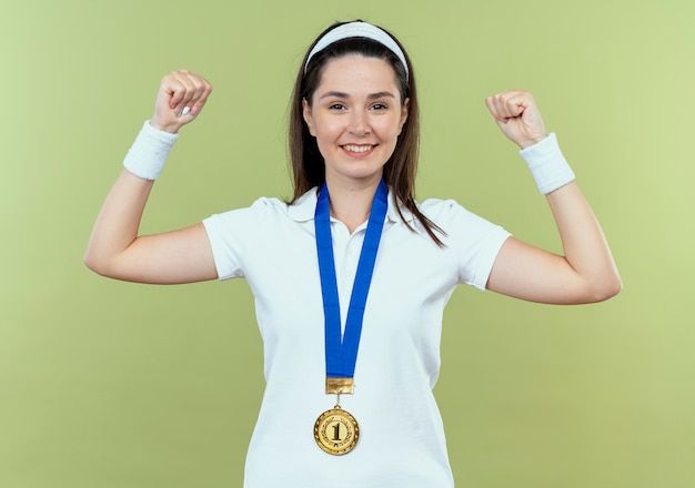 Bezpłatne zdjęcie młoda kobieta fitness w opasce ze złotym medalem na szyi, podnosząc pięść, patrząc pewnie z szczęśliwą twarzą uśmiechniętą stojącą na jasnym tle