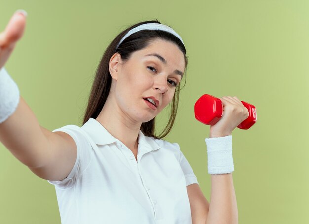 Młoda kobieta fitness w pałąku z hantlami, patrząc na kamery z pewną siebie wypowiedzi, stojąc na jasnym tle