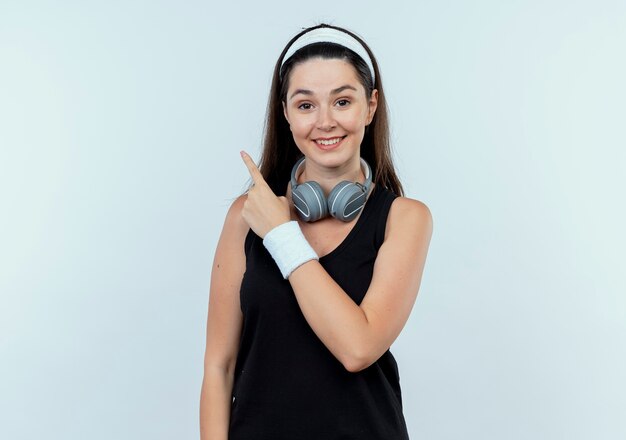 młoda kobieta fitness w pałąku ze słuchawkami, wskazując palcem w bok, uśmiechając się z radosną twarzą stojącą nad białą ścianą