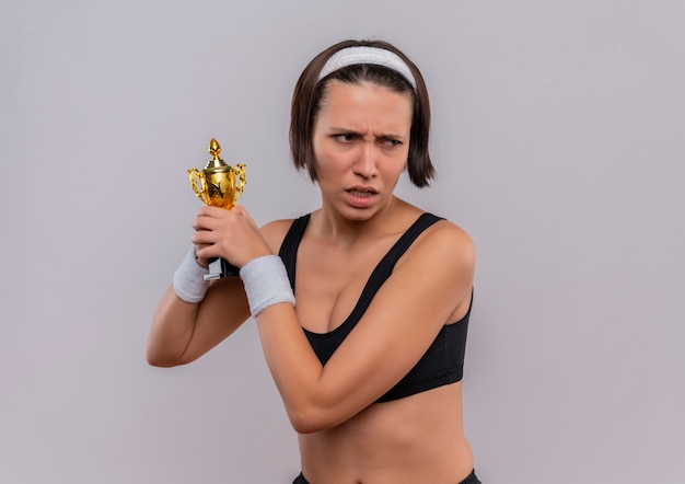 Bezpłatne zdjęcie młoda kobieta fitness w sportowej, trzymając jej trofeum patrząc na bok z marszczącą brwią twarz stojącą na białej ścianie