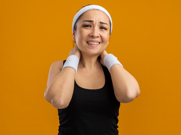 Młoda kobieta fitness z opaską na głowę i opaskami dotykającymi jej szyi, wyglądająca źle, czując ból, stojąc nad pomarańczową ścianą
