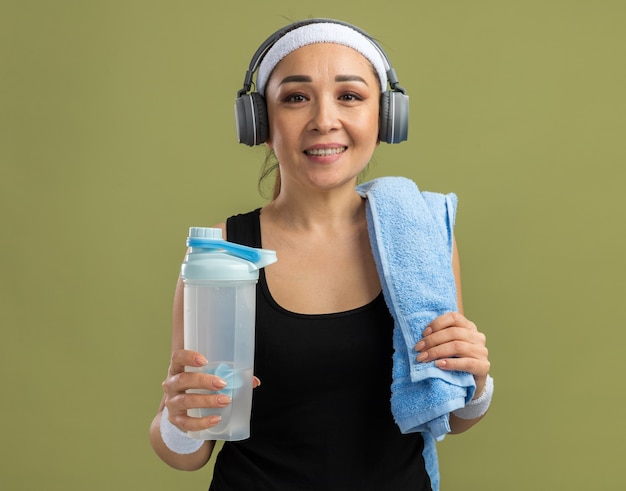 Młoda kobieta fitness z opaską na głowę i słuchawkami z ręcznikiem na ramieniu trzymająca butelkę wody z uśmiechem na twarzy stojącej nad zieloną ścianą
