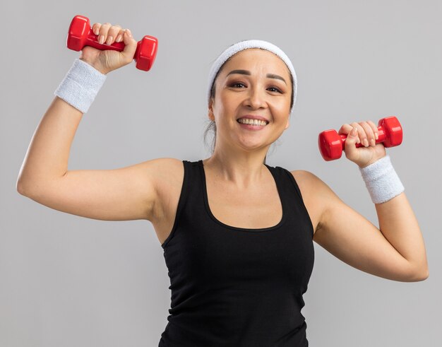 Młoda kobieta fitness z opaską na głowę z hantlami robi ćwiczenia, wygląda na zmęczoną i pewnie uśmiechniętą, stojąc nad białą ścianą