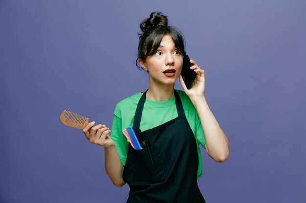 Młoda kobieta fryzjerka nosząca fartuch, trzymająca szczotkę do włosów, rozmawiająca przez telefon komórkowy, wyglądająca na zaskoczoną, stojącą na niebieskim tle