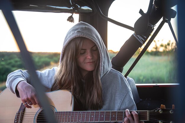 Młoda kobieta gra na gitarze akustycznej w bagażniku samochodu w naturze