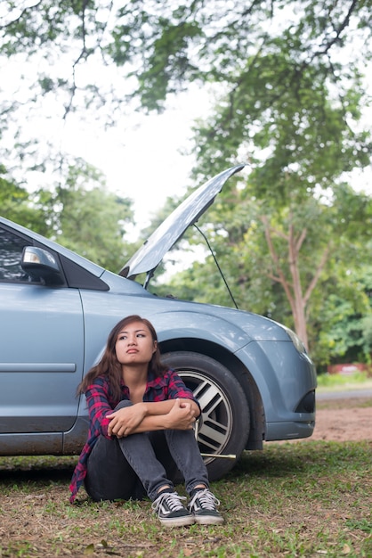 Młoda kobieta hipster czeka na pomocy drogowej po jej samochód rozkłada się z boku drogi siedzącej przeciwko jej samochodu.
