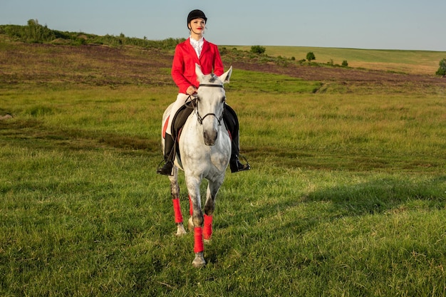 Młoda kobieta jeździec, ubrana w czerwone redingote i białe bryczesy, z koniem w wieczornym świetle zachodzącego słońca. Fotografia plenerowa w lifestylowym nastroju