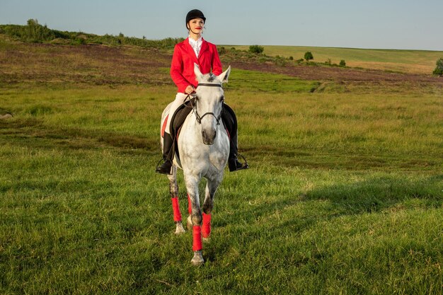 Młoda kobieta jeździec, ubrana w czerwone redingote i białe bryczesy, z koniem w wieczornym świetle zachodzącego słońca. Fotografia plenerowa w lifestylowym nastroju