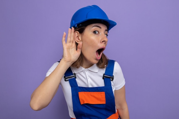 Bezpłatne zdjęcie młoda kobieta konstruktora w mundurze budowlanym i kasku wygląda na zdziwioną i zaskoczoną ręką nad uchem, próbując słuchać plotek stojących nad fioletową ścianą