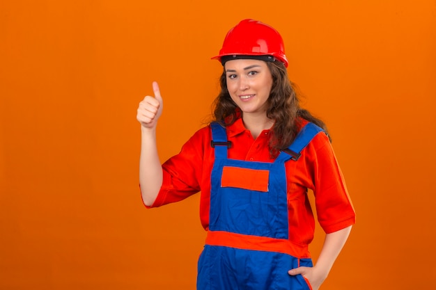 Młoda kobieta konstruktora w mundurze budowy i kask ochronny uśmiechnięty pokazując kciuki do góry na pojedyncze pomarańczowe ściany