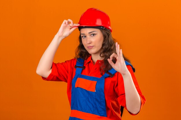 Bezpłatne zdjęcie młoda kobieta konstruktora w mundurze konstrukcyjnym i kasku ochronnym z uśmiechem dotykając kasku i robi ok znak nad izolowaną pomarańczową ścianą