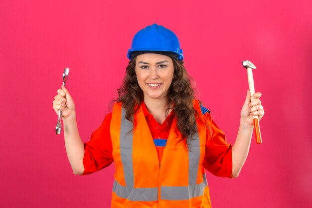 Młoda kobieta konstruktora w mundurze konstrukcyjnym i kasku stojącym z kluczem i młotkiem w uniesionych rękach uśmiechnięty przyjazny na izolowanych różowej ścianie