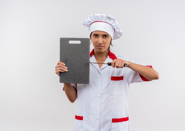 młoda kobieta kucharz ubrana w mundur szefa kuchni trzymając deskę do krojenia i nóż na izolowanych białej ścianie z miejsca na kopię