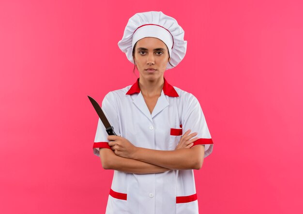 młoda kobieta kucharz ubrana w mundur szefa kuchni trzymając nóż i skrzyżowanie rąk na izolowanych różowej ścianie z miejsca na kopię